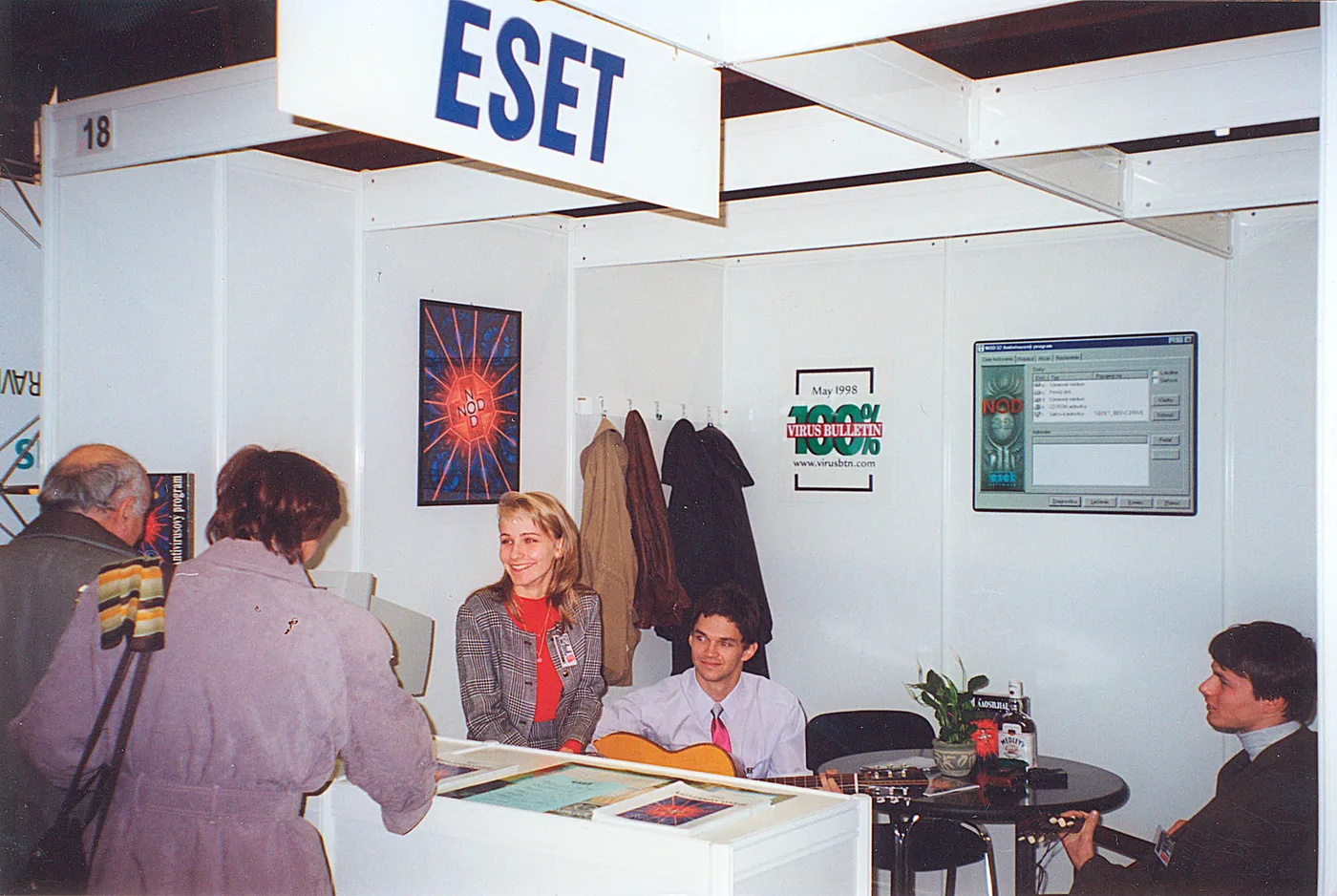 Stand de ESET en una feria, con un cartel colgante, una pantalla mostrando NOD32 y un póster del premio Virus Bulletin 100% de mayo de 1998; un joven toca la guitarra sentado junto al mostrador.