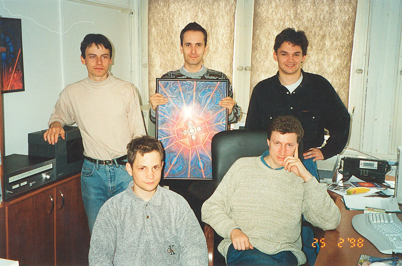 Cinco personas posando en una oficina modesta; la del centro sostiene un póster enmarcado con el símbolo de NOD. La cámara estampó la fecha de febrero de 1998 sobre la imagen.