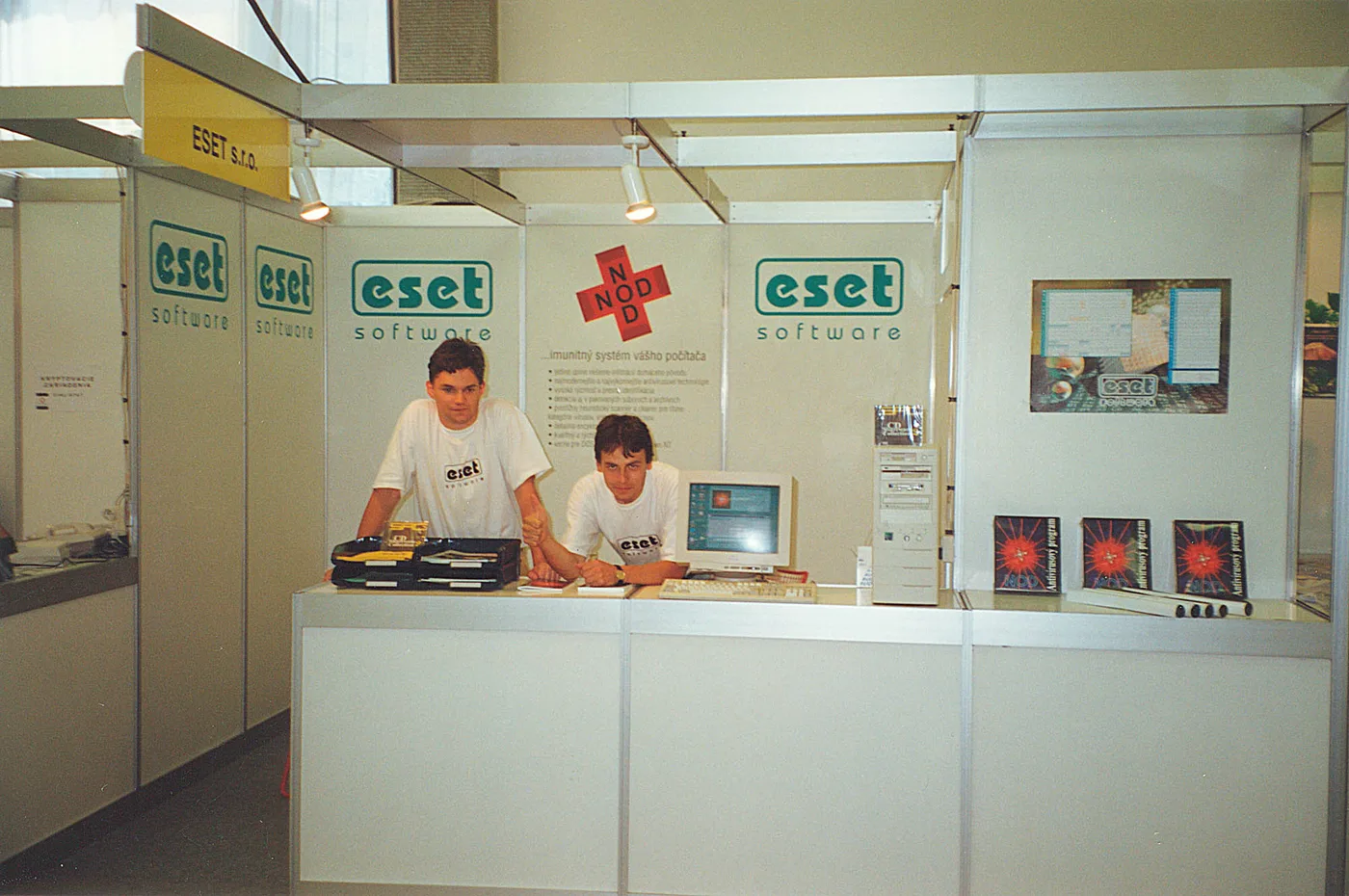 Stand de feria con paneles de «eset software» en verde y una cruz roja con la palabra NOD; dos jóvenes con remeras del logotipo atienden detrás del mostrador.