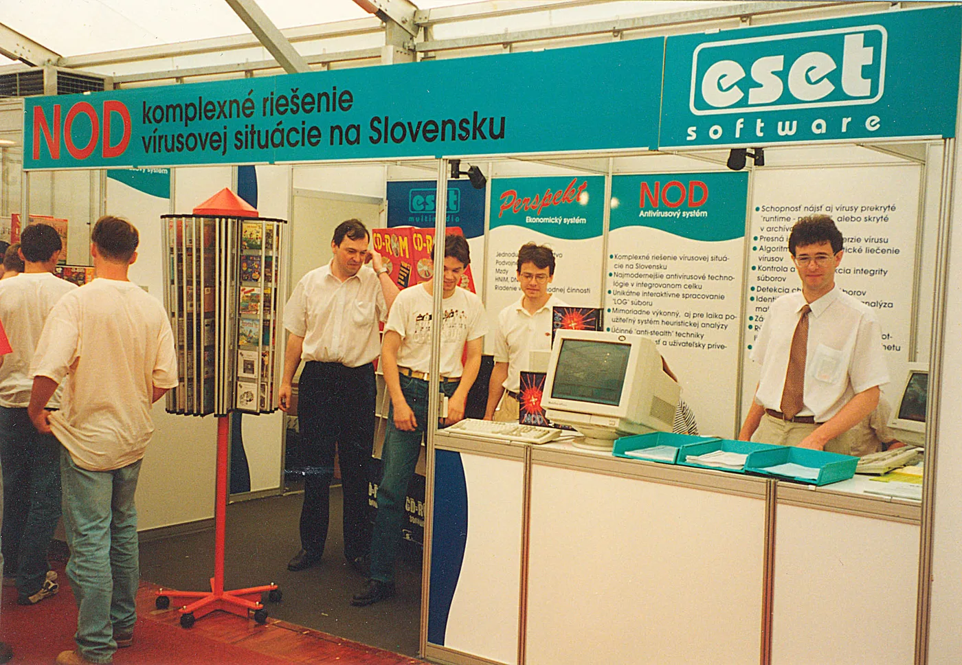 Stand de feria con una cartelera turquesa en eslovaco y el logotipo de eset software; varias personas conversan junto a un monitor de tubo.