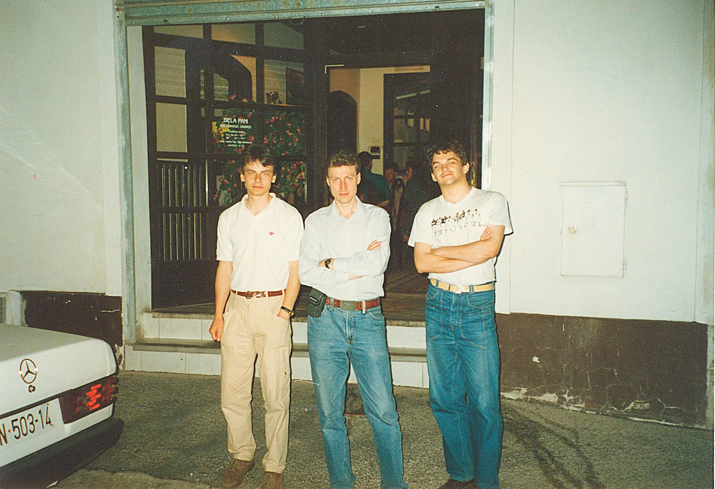 Tres hombres jóvenes posando de brazos cruzados en una calle, frente a la vidriera de un restaurante, junto a un auto blanco.